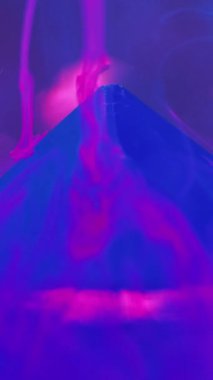 Dikey video. Su dumanı geçmişi. Mürekkep girdabı. Belirsizlik etkisi. Neon mavisi duman illüzyonu boya, mor sanatta taş piramitle sıvı maddede hareket eder..
