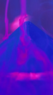 Dikey video. Su dumanı geçmişi. Mürekkep girdabı. Belirsizlik etkisi. Neon mavisi duman illüzyonu boya, mor sanatta taş piramitle sıvı maddede hareket eder..