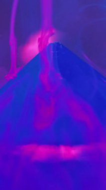 Dikey video. Su dumanı geçmişi. Mürekkep girdabı. Belirsizlik etkisi. Neon mavisi duman illüzyonu boya, mor sanatta taş piramitle sıvı maddede hareket eder..