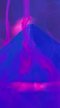 Dikey video. Su dumanı geçmişi. Mürekkep girdabı. Belirsizlik etkisi. Neon mavisi duman illüzyonu boya, mor sanatta taş piramitle sıvı maddede hareket eder..