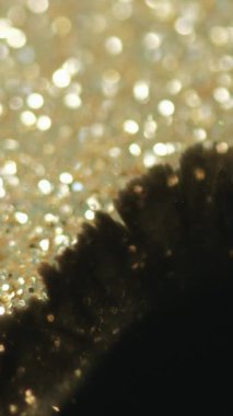 Dikey video. Mürekkep damlası. Boya serpintisi. Parlak altın parıltılı parçacıklar üzerindeki siyah ıslak çamur lekesi lekesi hareketi soyut arkaplan dokusundaki bokeh ışık dokusunu ortaya çıkarır.