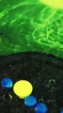 Dikey video. Boya baloncukları. Yağlı sıvı sıçraması. Odaklanmamış neon yeşil sarı renk akrilik mürekkep boyalı su damlacıkları koyu siyah soyut sanat arka planında yüzüyor..