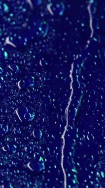 Dikey video. Yoğunlaştırılmış akış. Akıntıyı bırak Pürüzsüz mavi yüzeyde şeffaf çiy taneleri. Islak dokuda yansımaları olan sulu koyu arkaplan.