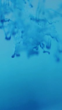 Dikey video. Renkli su damlası. Boya sıçraması. Odaklanmamış sıvı soyut sanat arka planında mavi sıvı damlacıklar dumanlı bulutlar akıtır.