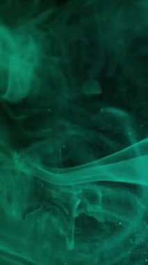 Dikey video. Duman patlaması. Duman spreyi. Yeşil turkuaz boya akıntıları karanlık sularda yayılıyor. Parlak odaklı soyut gece dokusu arkaplanı.