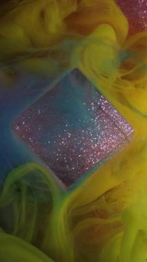 Dikey video. Renkleri karıştırıyorum. Boya dökülmüş. Parlak mavi duman dumanı simli küp mürekkep dokusu mor parıldayan sanat arka planında yüzüyor..