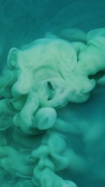 Dikey video. Harman boyası. Akrilik rengi. Parlak sıvı parçacıkların dumanının içine dökülen çözünmüş yeşil mavi bulut soyut arkaplan üzerine düşer.