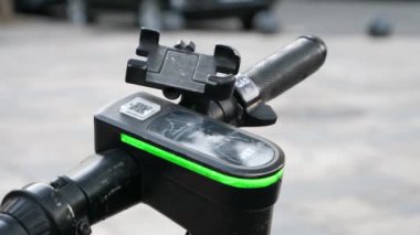Elektrikli scooter 'ın yakın çekim aşınmış direksiyonu yanıp sönen yeşil LED şerit lambası, QR kodu ve cep telefonu kablosu ile. Şarj edildi ya da e-scooter şarj edildi. Tiflis, Gürcistan - 22 Kasım 2024.