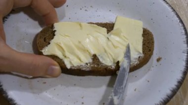 Eller tereyağını beyaz bir tabakta siyah bir ekmek dilimine kolayca yayıyor, günlük basit bir mutfak görevini detaylıca anlatıyor, yüksek açılı yakın plan bir görüntü..