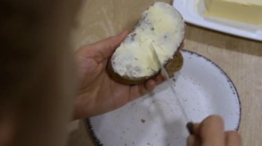 Kadın, tereyağını beyaz bir tabakta, siyah bir ekmek diliminde, bıçakla düzgünce yayıyor, günlük basit bir mutfak görevini, omuz manzarası üzerinden detaylandırıyor..