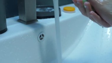 Bir çocuk, ellerini banyo lavabosunda sabun ve suyla iyice yıkayarak el hijyenini takip eder. Ellerinde yakın bir sol görüş, bir musluk ve beyaz bir lavabo..