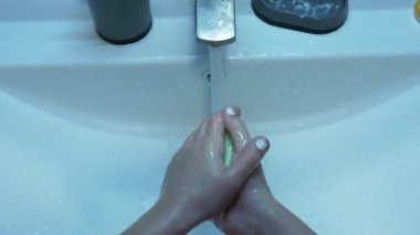 Kız ellerini banyo lavabosunda sabun ve suyla iyice yıkayarak doğru dürüst el hijyenini takip ediyor. Yüksek açılı el kamerası, musluk ve beyaz bir lavabo..