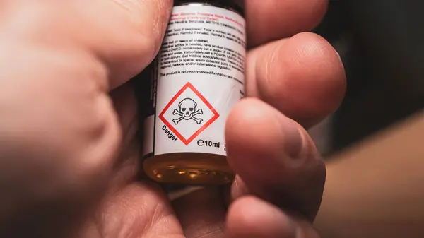 Hand holding a small bottle of e-cigarette nicotine liquid, displaying a danger skull sign, indicating potential health risks associated with vaping.