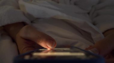 Bir kadının eli, beyaz bir bornoz giyip yatakta yatarken, akıllı telefon ekranında sosyal medyada gezinirken yakın plan görüntüsü..