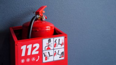 Parlak kırmızı bir yangın söndürücü kırmızı bir kutunun içinde saklanıyor. Acil durum numarası 112 ve kullanım talimatları yazıyor..