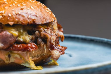 Ekmeğin üzerinde susamlı hamburger ve mavi bir tabakta dinlenen pastırma. Yakın plan çekimde hamburger malzemelerinin ayrıntıları vurgulanıyor..