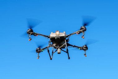 Açık mavi gökyüzüne karşı havada uçan böcek ilacı püskürtücü bir dron. İnsansız hava aracının dört pervanesi, bir sprey tankı var ve sprey ilaçlama uygulaması için enjektörlerle donatılmış..