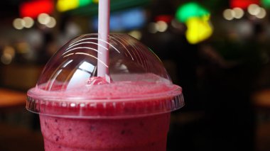 Plastik bardakta, parlak bir kafe ortamında pipetle pembe bir smoothie 'nin yakın plan çekimi. Smoothie 'nin üstü şeffaf plastik kubbe kapağıyla kaplı..