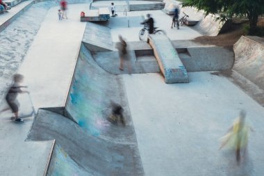 Beton bir kaykay parkında kayan ve bisiklete binen insanların bulanık siluetleri rampa ve raylara karşı dinamik hareketlerini vurguluyor, uzun pozlu çekim.