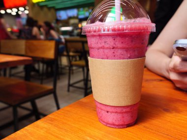 A pink smoothie in a plastic cup sits on a wooden table in a Cafe. The smoothie cup has an empty paper cup sleeve on it. Carton cup sleeve mockup. clipart