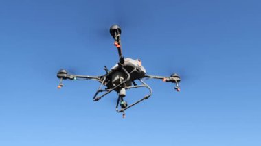 Açık mavi gökyüzüne karşı havada uçan böcek ilacı püskürtücü bir dron. İnsansız hava aracının dört pervanesi, bir sprey tankı var ve sprey ilaçlama uygulaması için enjektörlerle donatılmış..