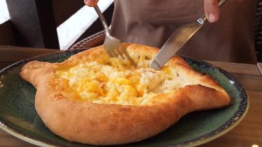Bir insan, geleneksel Gürcistan mutfağının altını çizerek, taze pişmiş bir Adjarian Khachapuri restoranında peynirle yumurtayı karıştırmak için çatal ve bıçak kullanır..
