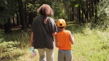 Bir anne ve oğlu, ağaçlar ve yeşilliklerle çevrili bir orman yolunda el ele yürürler ve doğada sıcak bir günün tadını çıkarırlar..