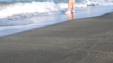 Kumlu sahil boyunca yalınayak yürüyen kişi dalgalar yavaşça içeri giriyor. Okyanus dalgaları kıyıda süzülürken ayakların yumuşak kumda geziniyor..