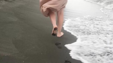Beyaz bir kadın kumlu bir plajda çıplak ayakla yürüyor, arkasında ayak izleri bırakıyor, ve soğuk bir günde bej bir elbise giyiyor, orta boy bir atış yapıyor..