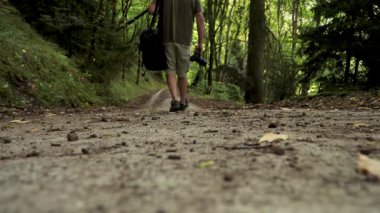 Vahşi yaşam fotoğrafçısı, 4K yavaş çekim ile yemyeşil bir yaz ormanını keşfediyor. Bir gezgin, uzun bir yolda, canlı bir ormanda elinde bir kamera ve çantayla dolaşır..