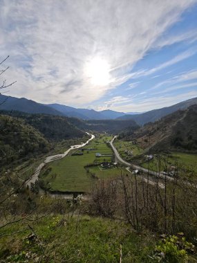 Parlak bir güneş ve bulutlu bir gökyüzü altında kıvrılan bir nehir, evler, yollar ve dağların manzarası.