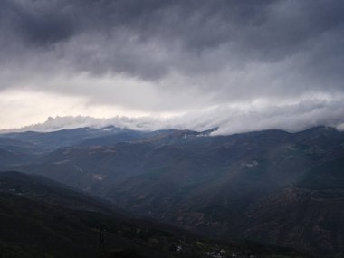Kara fırtına bulutları bir dağ sırasının üzerinde toplanıyor, aşağıdaki vadide dramatik ve kasvetli bir atmosfer yaratıyorlar.
