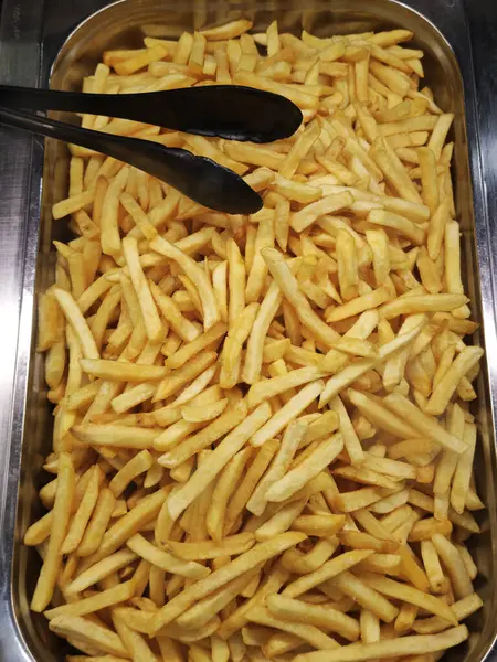 Metal tepsi büyük miktarda altın patates kızartmasıyla doludur. Tepesinde siyah maşayla servis edilmeye hazırdır.