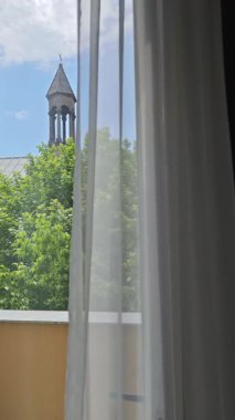 Güneş ışığı, beyaz perdelerin arasından bir otel odasının penceresinden görülen bir kilise kulesini aydınlatır.