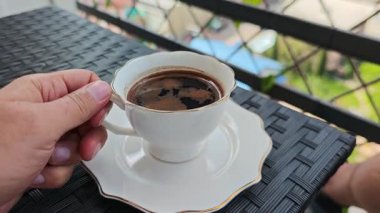 Elle bir fincan kahveyi güneşli bir balkondan kaldırır ve dışarı çıkarak rahatlatıcı bir an yaşatır.