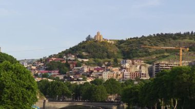 Mtkvari nehri ve yemyeşil ağaçlar, Tiflis renkli evleri ve Aziz Nicholas Kilisesi 'nin bir tepenin üzerinde durduğu manzarayı oluşturur. Tiflis, Gürcistan - 26 Mayıs 2025.