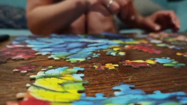 Beyaz ojeli kadın elleri yapboz parçalarını ahşap bir masaya yerleştiriyor ve renkli bir resim oluşturuyor.