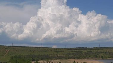 Ağaçlar ve elektrik kuleleri olan yeşil bir arazide beyaz kümülüs bulutları büyüyor.