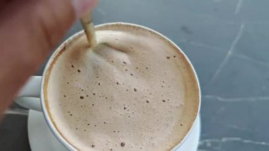 Profesyonel barista kremalı kapuçino köpüğünü gümüş kaşıkla nazikçe karıştırarak kafe reyonundaki beyaz seramik bardakta köpüklü dokular oluşturuyor.