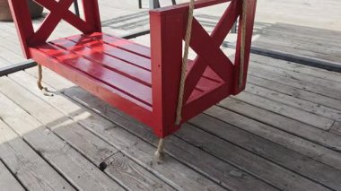 Canlı kırmızı ahşap salıncak bankı halatlarla asılmış, güneşli açık hava ahşap terasında dinleniyor, minimalist bahçe tasarımını vurguluyor.