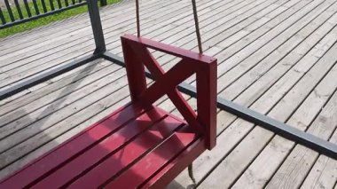 Klasik kırmızı ahşap salıncak. Sundurmada yavaşça hareket ediyor. Ritmik ve nostaljik hareketlerle huzurlu bir yaz atmosferi yaratıyor.