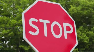 Kırmızı dur işareti yeşil ağaçlara doğru yavaşça sallanıyor, yol kenarı hareketini parlak gün ışığında, sakin, doğal arka planda ve net trafik düzenlemeleri görsel olarak yakalıyor.