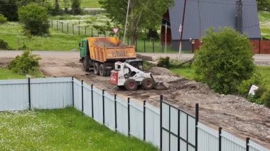 Ağır makineler yol yapımında kullanılıyor, endüstriyel fren dümeni yükleyicisi ve çöp kamyonu birlikte çalışıyor, toprağı boşaltıyor ve kırsal alanda hareket ettiriyor. Tsemi, Gürcistan - 6 Haziran 2025.