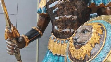 Elinde kılıç ve kalkan tutan bir Romalı askerin bronz heykeli modern bir binaya gururla karşı duruyor.