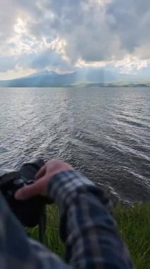 Kamera ayarlarını ayarlayan fotoğrafçı, dalgalı göl ve bulutlu gökyüzünün altındaki görkemli dağın çarpıcı görüntüsünü yakalıyor.