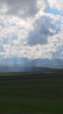Verdant Vadisi karlı dağların altında yayılıyor, süzülen bulutlarla süzülen yeşil manzara parlak güneş ışığı altında yumuşak gölgeler bırakıyor.