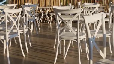 Beyaz sandalyeler ve masalar düzenli bir şekilde tekrar tekrar sıralanır, güneş ışığıyla yıkanır, dingin ve davetkar bir atmosfer yaratır.