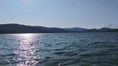 Güneş ışığı, arkaplanda açık mavi gökyüzü ve dağlarla dalgalanan su yüzeyini yansıtır.