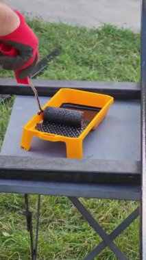 İnşaat işçisi metal ızgaraya siyah boya uyguluyor. Tekerlek kullanıyor, dış bakım sırasında koruyucu kırmızı eldiven takıyor.