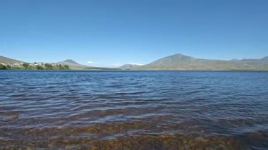 Sakin su yüzeyinde parlayan güneş ışığı, dingin arkaplanda yükselen dağ siluetleri, manzaralı göl ortamının huzurlu doğal güzelliğini yansıtıyor.